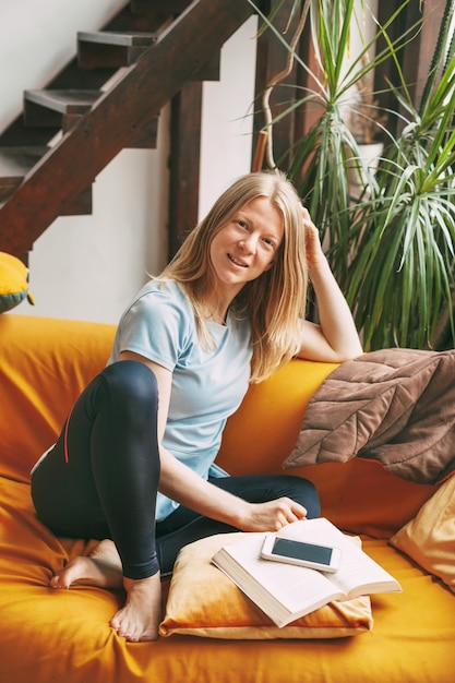 Cute girl sitting on the couch and smiling, next to an open book and phone