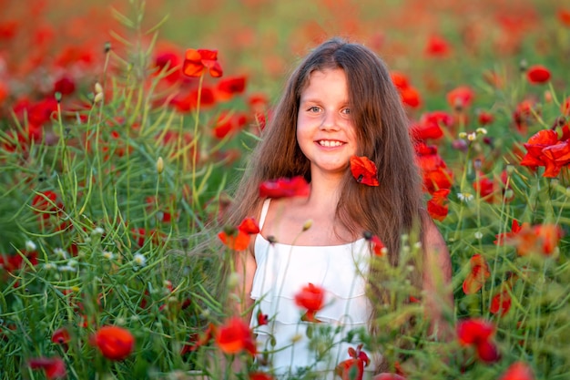 Cute girl wearing white dress in summer blooming poppy field walking under evening sun