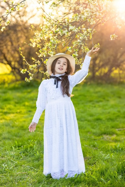 A cute girl in a white dress and a hat under flowering trees in the garden