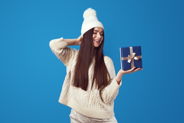 Cute girl in white sweater and hat holding wrapped blue gift box for