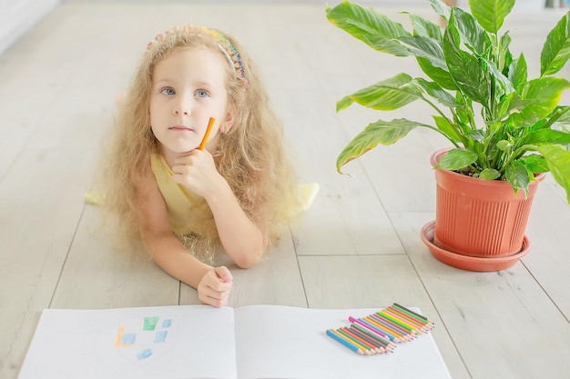 a cute girl with blue eyes draws a houseplant next to her in an album with colored pencils