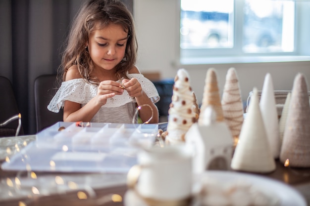 Cute girl with curly hair is handcrafting and decorating paper cone Christmas Tree with buttons, yarn and fairy lights. 