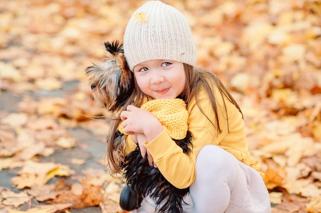 Cute girl with a fallen yellow leaf on a hat, gently hugs her pet dog in an autumn park against a background of yellow fallen leaves