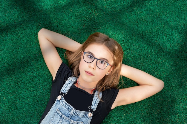 Cute girl with glasses looking at camera lying on the grass with her hands on the back of her head