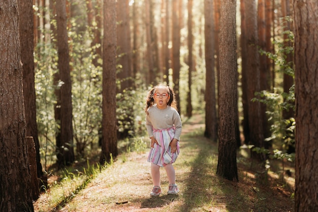 Cute girl with glasses makes a funny grimace and shows tongue in nature in summer