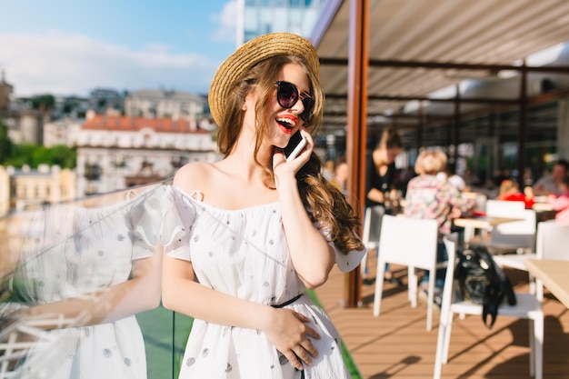 Cute girl with long hair in sunglasses is standing on the terrace. She wears a white dress with bare shoulders, red lipstick and hat . She is speaking on phone and looking to the side