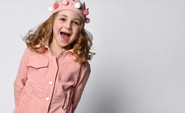 Cute girl with a plush crown standing upright in a dusty rose cotton summer jumpsuit with