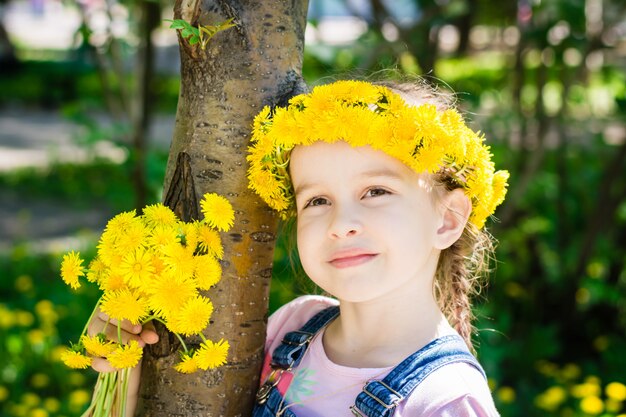 Cute girl in a wreath of dandelions and with a bouquet of dandelions in her hand for a walk in the park