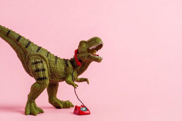 Cute green toy dinosaur speaking on retro phone on pink background Copy space