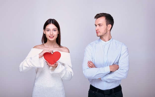 A cute guy with folded arms in a blue shirt looks at woman in white sweater who is holding a red cardboard heart