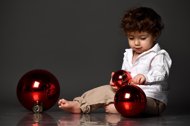 Cute handsome barefoot chubby baby boy in trendy outfit playing with Christmas toy bauble balls different size Isolated portrait shot on gray studio background Winter holiday Xmas eve concept