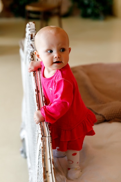 Cute happy blue-eyed 6-month-old girl in bright red dress at head of retro bed