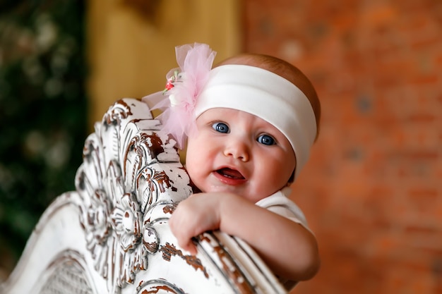 Cute happy blue-eyed 6-month-old girl in white headband at head of retro bed