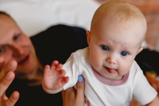 Cute happy blue-eyed 6-month-old girl in white outfit plays on bed with man's father