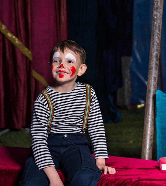 Photo cute happy boy with clown make up sitting on a stage before a representation while wearing striped long-sleeve shirt and braces