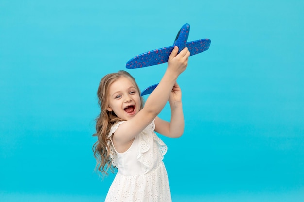 Cute happy little girl in a cotton white dress on a blue background with an airplane in her hands playing a place for text