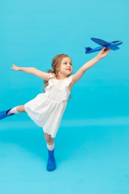 Cute happy little girl in a cotton white dress on a blue background with an airplane in her hands playing a place for text