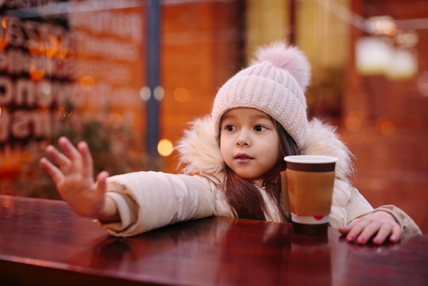 Cute happy little girl drinks hot tea in a street cafe