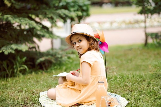 Cute happy little girl in a hat having fun in park meadow Child sitting on a blanket drawing picture and and look at camera Picnic on nature