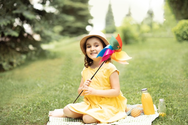 Cute happy little girl in a hat having fun in park meadow Child sitting on a blanket holding pinwheel and look at camera Picnic on nature