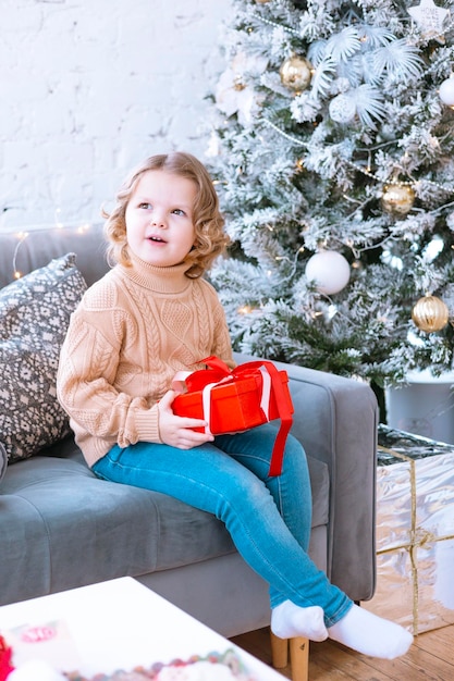 cute happy little girl with curls is sitting on chair near Christmas tree with gift box in her hands