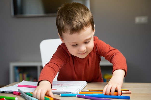 A cute happy little toddler boy of two years playing with markers in the album in the childrens room