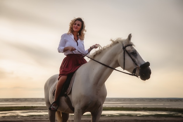 Cute happy young woman on horseback in summer beach by sea. Rider female drives her horse in nature on evening sunset light background. Concept of outdoor riding, sports and recreation. Copy space