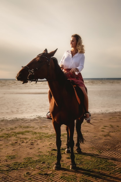 Cute happy young woman on horseback in summer beach by sea. Rider female drives her horse in nature on evening sunset light background. Concept of outdoor riding, sports and recreation. Copy space