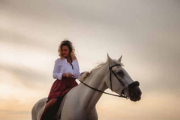 Cute happy young woman on horseback in summer beach by sea. Rider female drives her horse in nature on evening sunset light background. Concept of outdoor riding, sports and recreation. Copy space