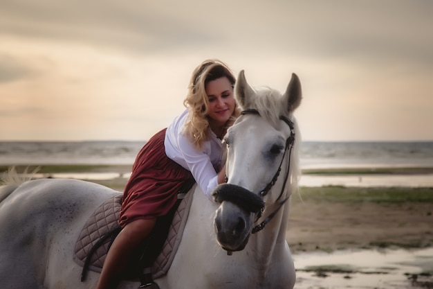 Cute happy young woman on horseback in summer beach by sea. Rider female drives her horse in nature on evening sunset light background. Concept of outdoor riding, sports and recreation. Copy space