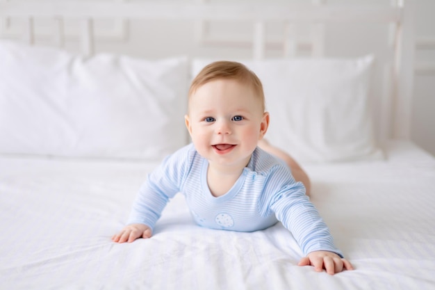 A cute healthy little baby is lying on a bed on white bedding at home in a blue bodysuit The kid looks at the camera smiles