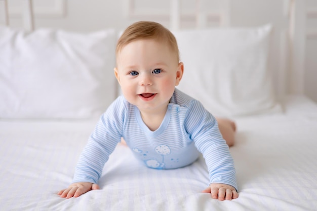 A cute healthy little baby is lying on a bed on white bedding at home in a blue bodysuit The kid looks at the camera smiles