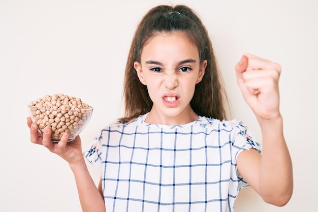 Cute hispanic child girl holding chickpeas bowl annoyed and frustrated shouting with anger yelling crazy with anger and hand raised