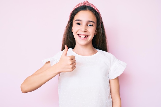 Cute hispanic child girl wearing casual clothes and diadem doing happy thumbs up gesture with hand. approving expression looking at the camera showing success.