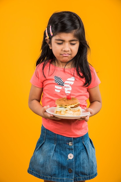 Cute Indian or Asian little girl eating tasty Burger, Sandwich or Pizza in a plate or box. Standing isolated over blue or yellow background.