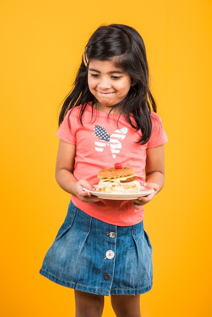 Cute Indian or Asian little girl eating tasty Burger, Sandwich or Pizza in a plate or box. Standing isolated over blue or yellow background.