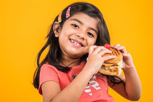 Cute Indian or Asian little girl eating tasty Burger, Sandwich or Pizza in a plate or box. Standing isolated over blue or yellow background.