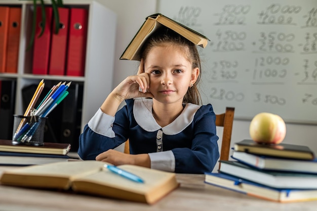 Cute industrious child is sitting at a table indoors The child is studying in the classroom on the background of the blackboard The girl is reading a book Back to school and happy times