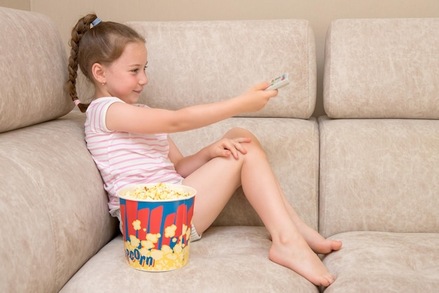 Cute joyful girl sits at home on the sofa with a huge glass of popcorn and switches TV channels