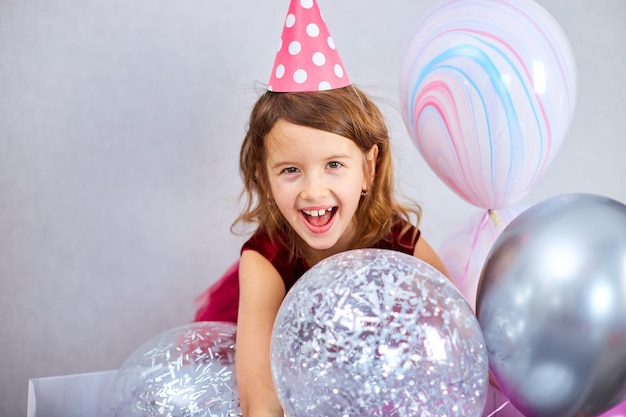 Cute Joyful little girl in pink dress and hat play with balloons at home birthday party streamers Happy birthday Celebrating