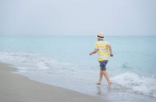 Cute kid boy running on the ocean or sea edge Child walking on the beach and playing with waves