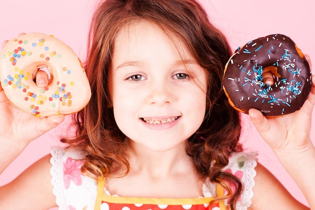 Cute kid girl holding tasty donut over pink background, Unhealthy eating