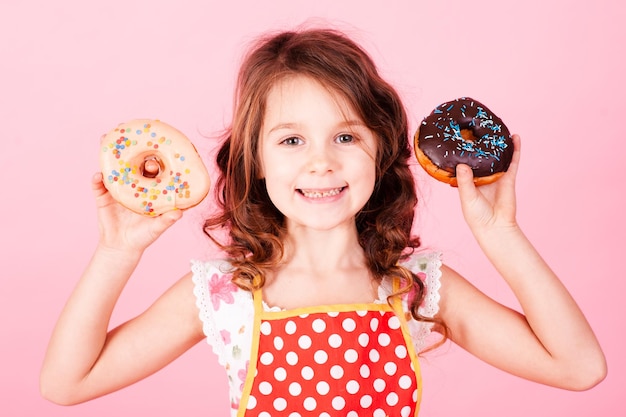 Cute kid girl holding tasty donut over pink background, Unhealthy eating