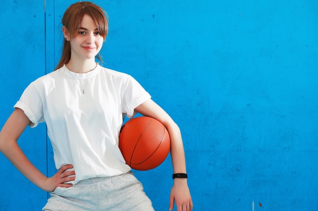 Photo a cute kid, girl is engaged in sport, she is looking aside while exercising with basketball. isolated onblue background. fitness, training, active lifestyle concept. horizontal shot. front view