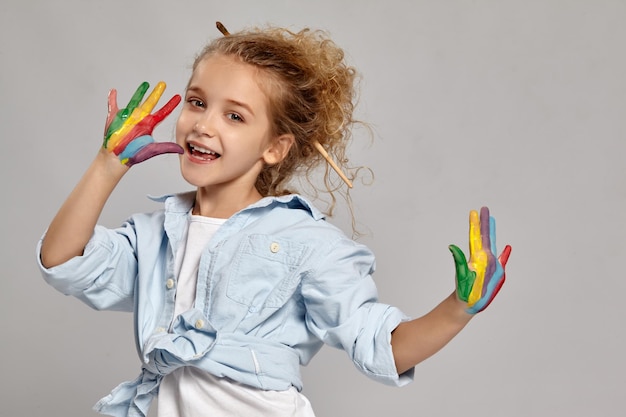 Cute kid having a brush in her chic curly blond hair, wearing in a blue shirt and white t-shirt. She is posing with a painted arms and smiling, on a gray background.