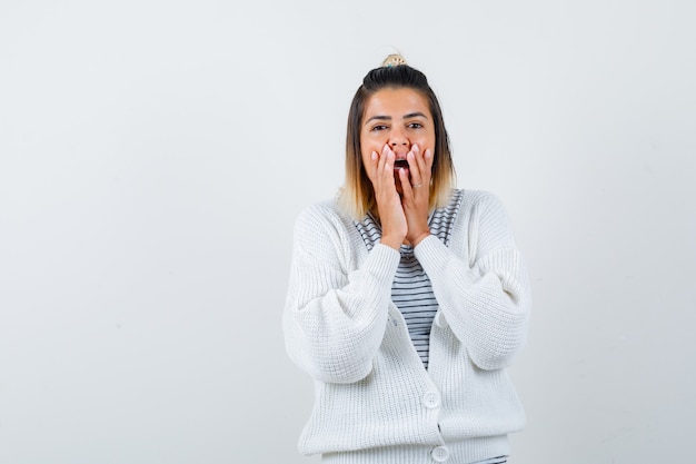 Cute lady in t-shirt, cardigan holding hands near open mouth and looking surprised