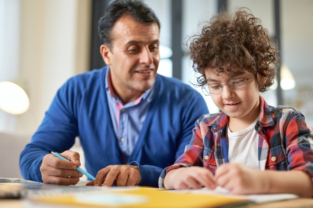 Photo cute latin boy wearing glasses looking focused while sitting at the desk together with his father and doing homework during remote learning at home. online education, homeschooling, parenthood