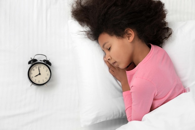 Cute little AfricanAmerican girl with alarm clock sleeping in bed top view