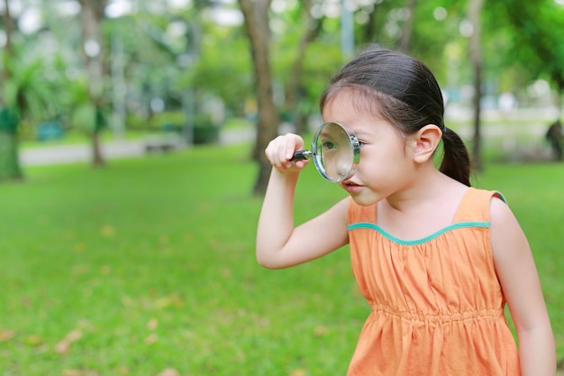 Cute little Asian child girl looking through magnifiying glass on at grass outdoors.