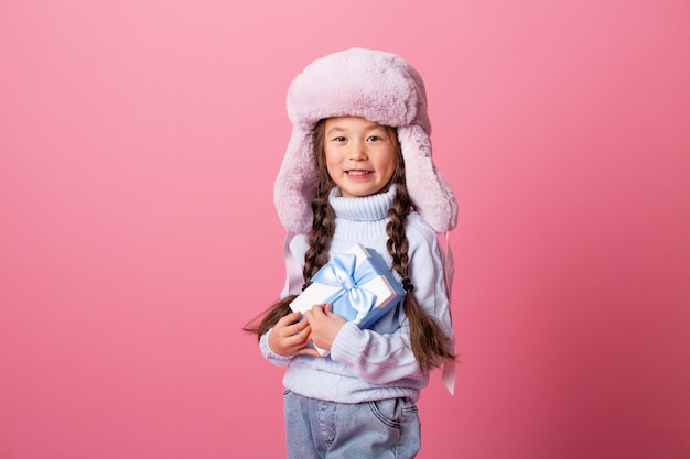 Cute little Asian girl in a winter hat and sweater holds a gift box. Christmas concept, text space